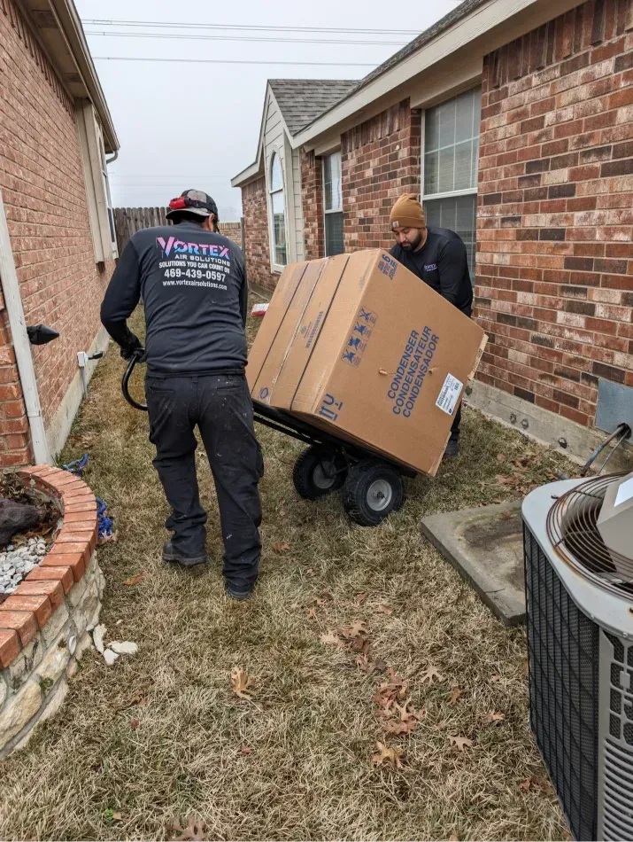 Two men moving a large cardboard box on a hand truck next to a brick house and an air conditioning unit.