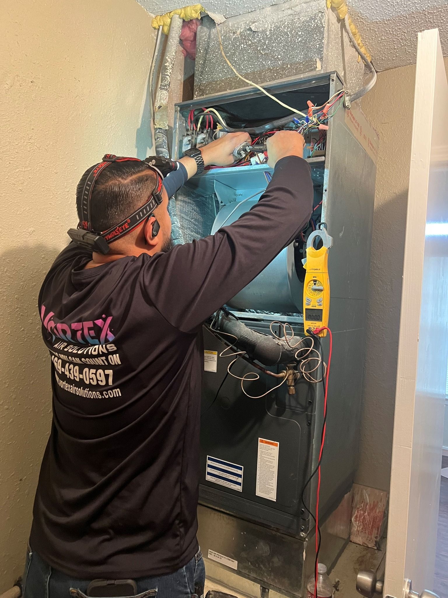 HVAC technician repairs an air handler unit. Wearing headlamp, dark shirt. Interior setting.