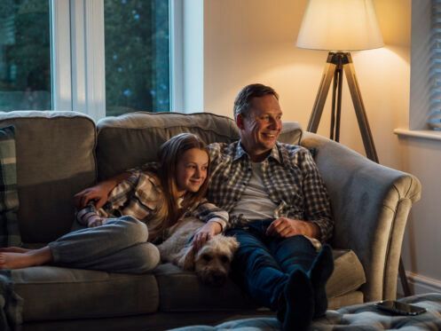A man and child smile while sitting on a couch next to a small dog in a living room.