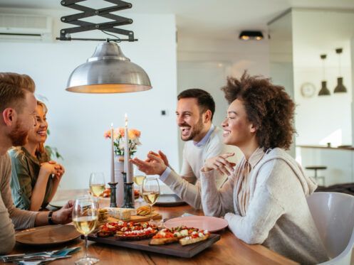 Friends laughing and dining at a wooden table set with food, drinks, and candles.