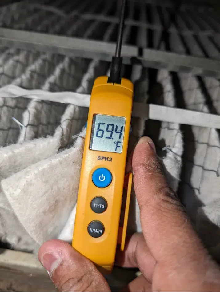A hand holding a yellow digital thermometer that displays 69.4°F, in front of a white filter in a metal frame.