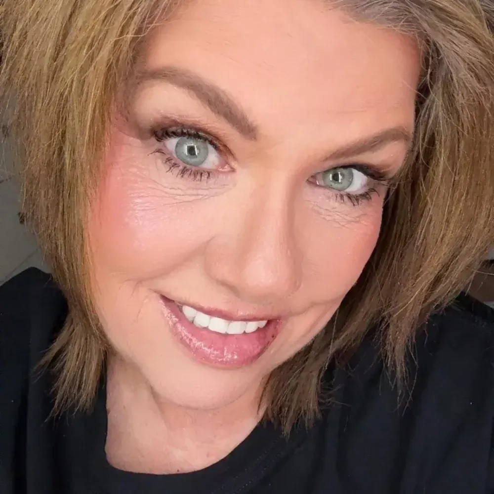 Woman with light brown hair smiles, eyes green. Wearing makeup, a black top.