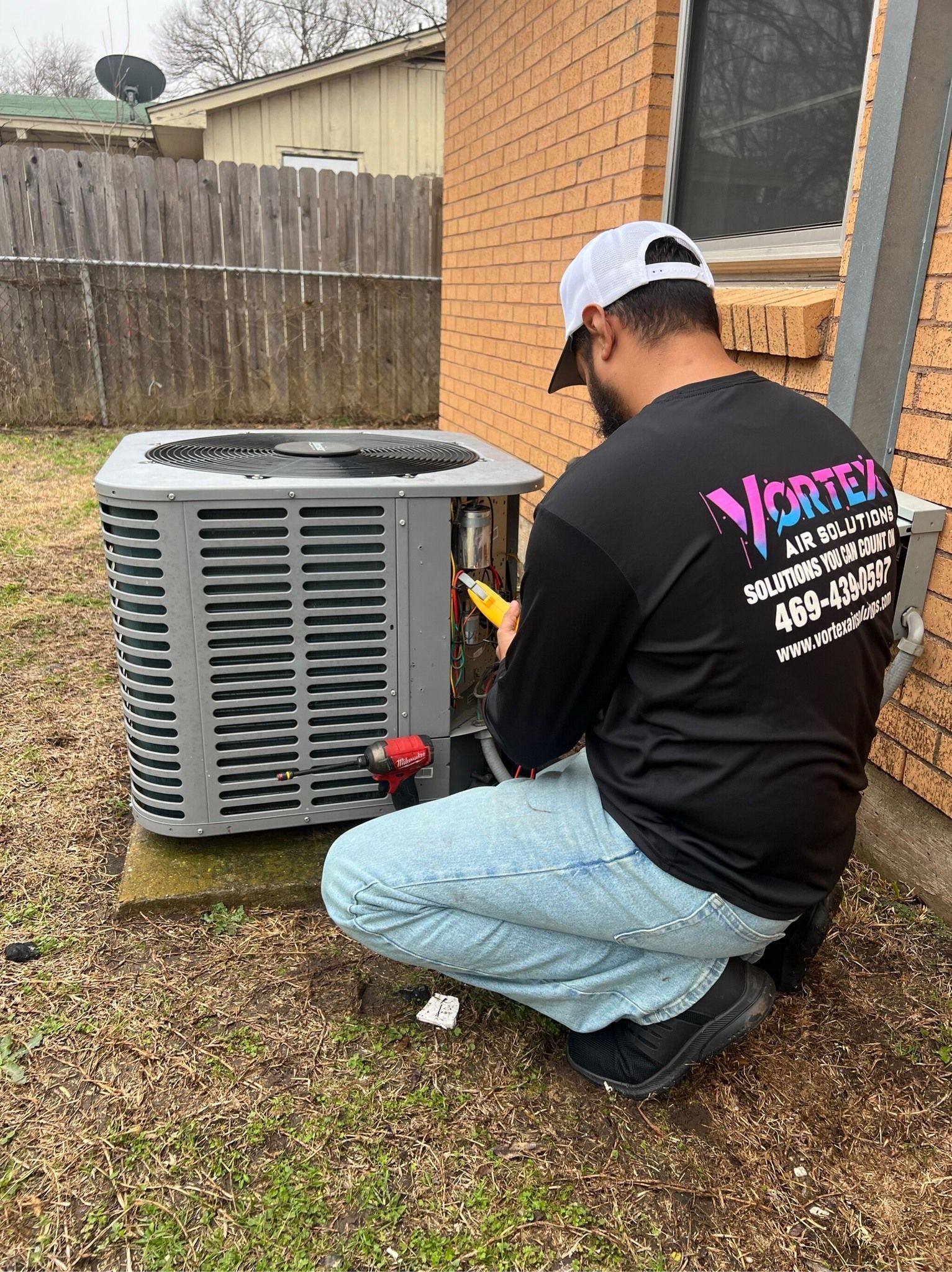 Man in a black shirt works on an AC unit outdoors. He kneels near the unit on grass, near a brick building.