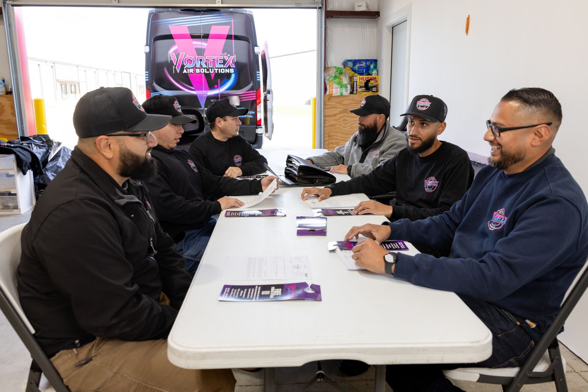 Person in black shirt sprays an AC unit with a green hose outdoors. Logo on shirt: Vortex Air Solutions.
