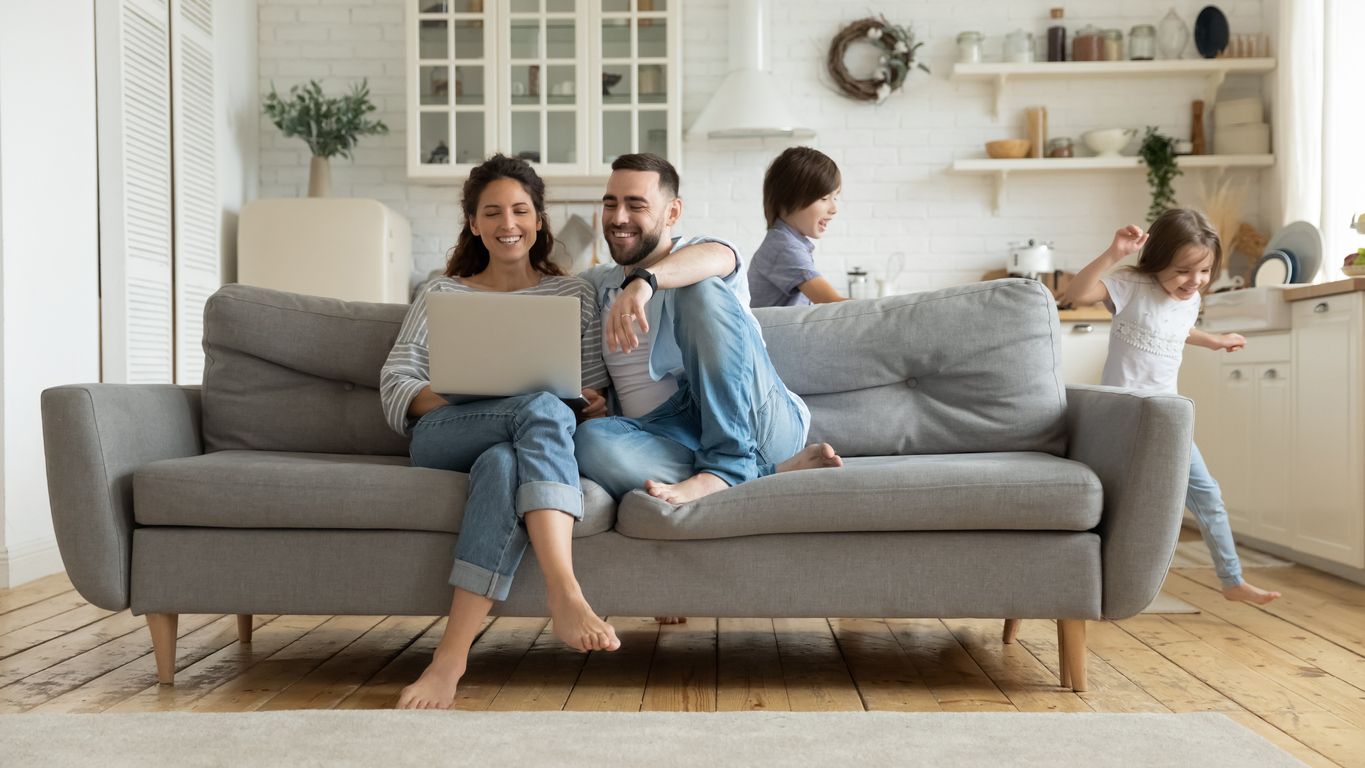 Family on a sofa with laptop, two children playing in kitchen. Smiling, light-filled room.