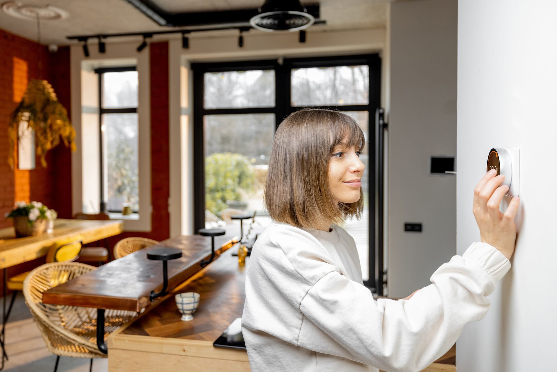 Woman adjusting a thermostat on a white wall in a modern dining room.