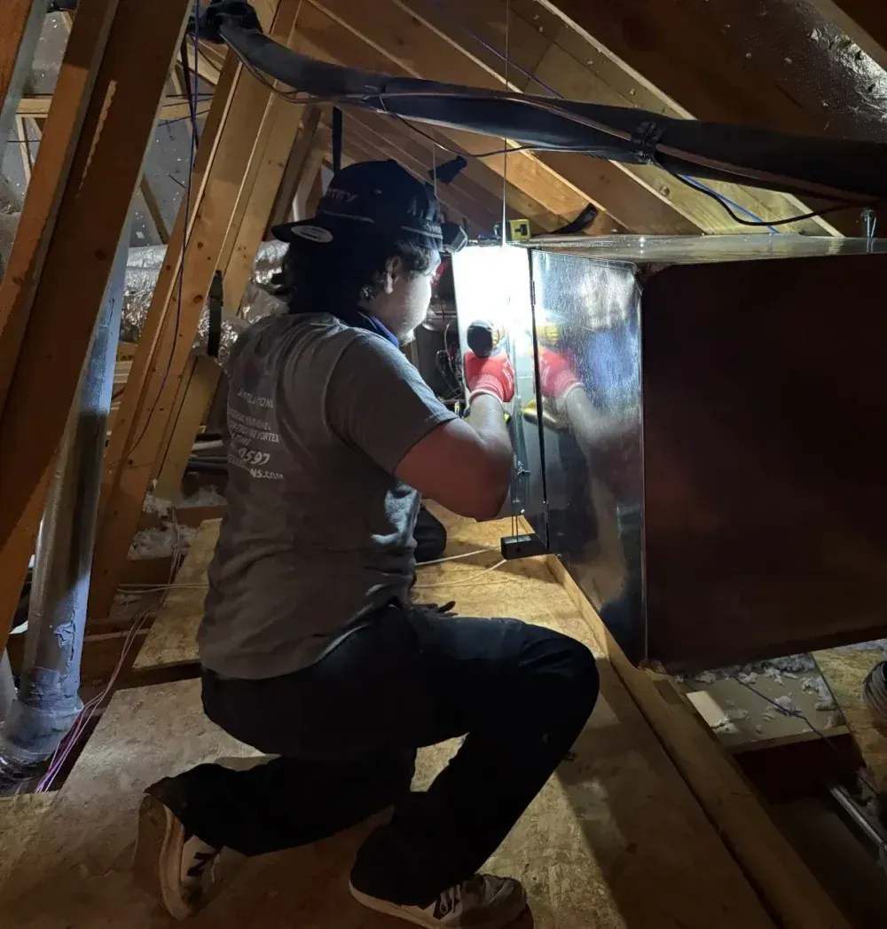 Person works on HVAC unit in attic, kneeling on wooden beams.