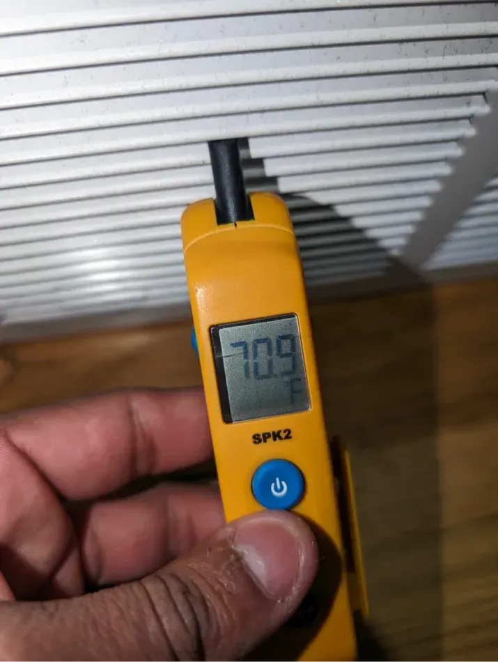 Hand holding a yellow thermometer measuring air temperature at a white vent. Reading is 70.9°F.