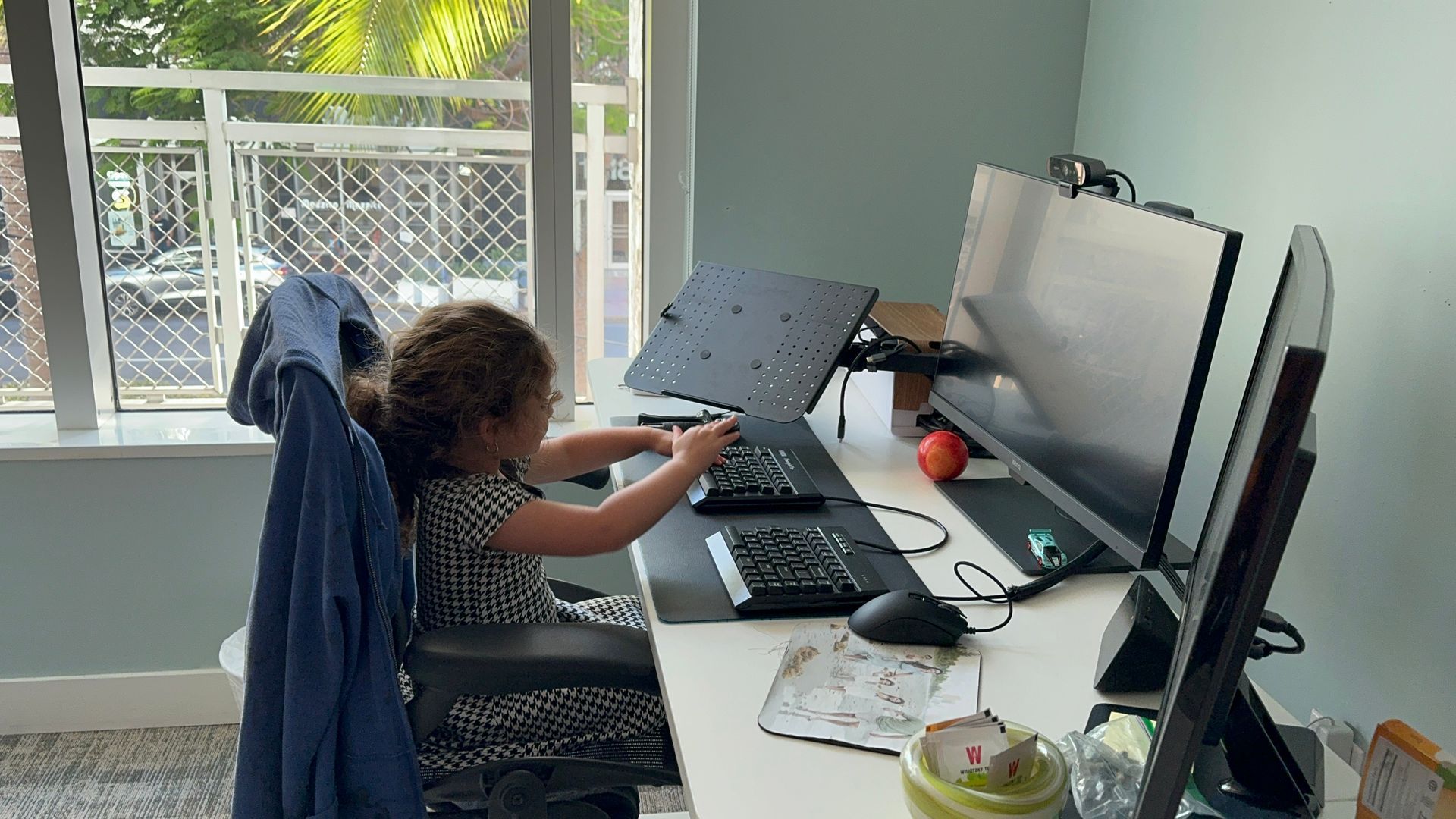 Young child at a desk, playing with computer, two monitors, window in background.