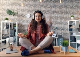 A woman is sitting on a desk in a lotus position with headphones on.