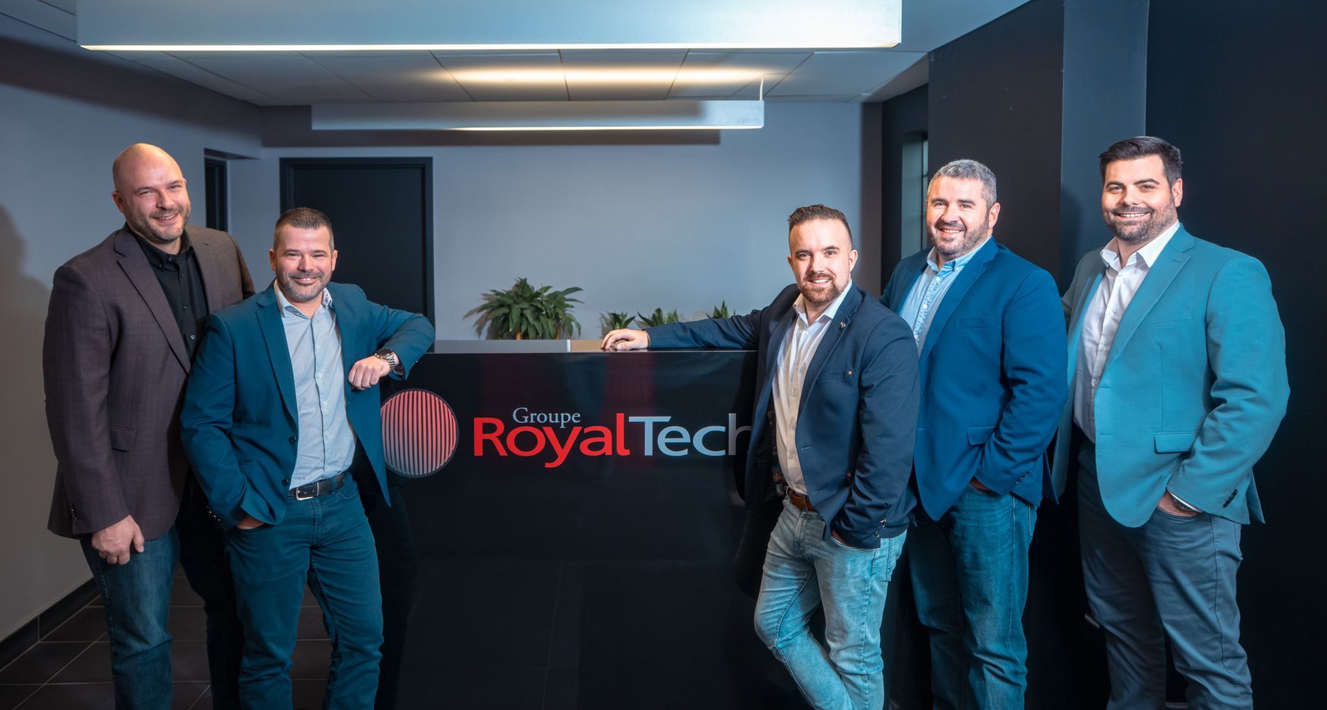 Cinq hommes se tiennent debout dans un bureau professionnel, appuyés contre un bureau noir portant le logo de la société RoyalTech.