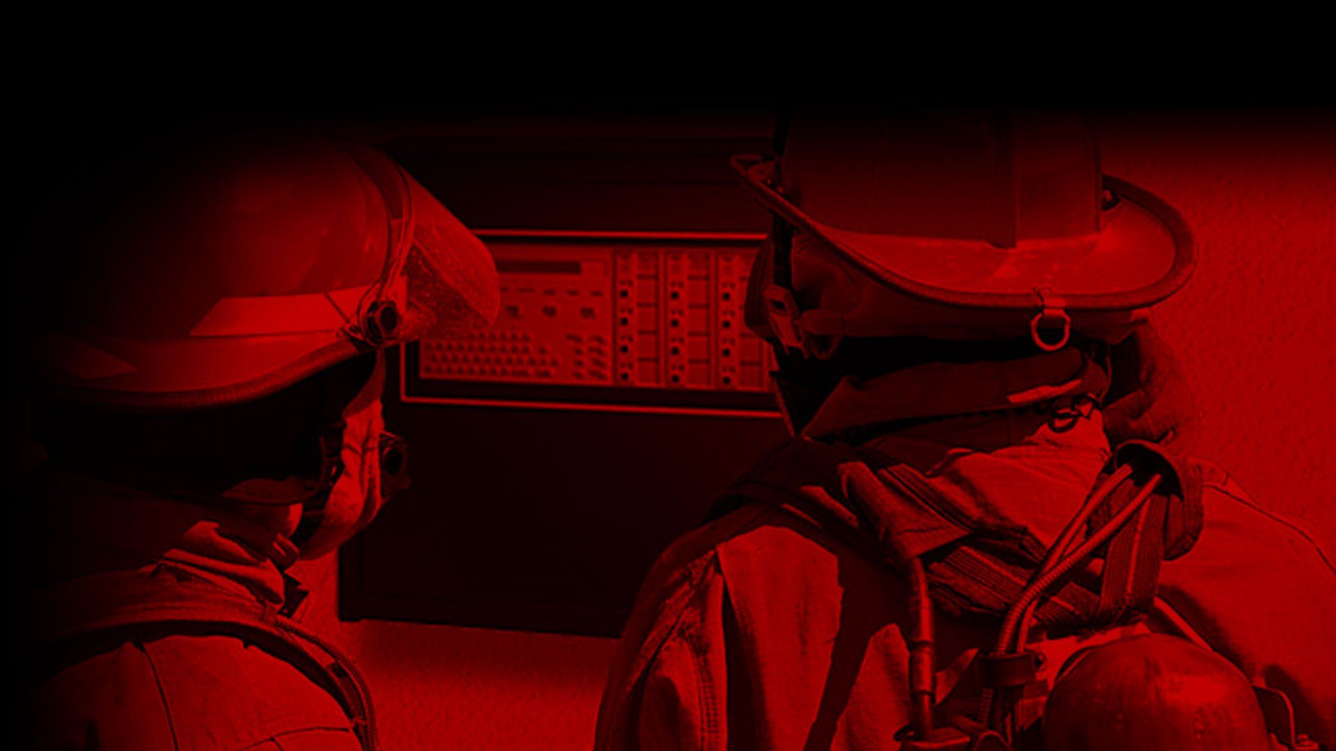 Two firefighters in helmets and gear, silhouetted in red light, facing a wall with a control panel.