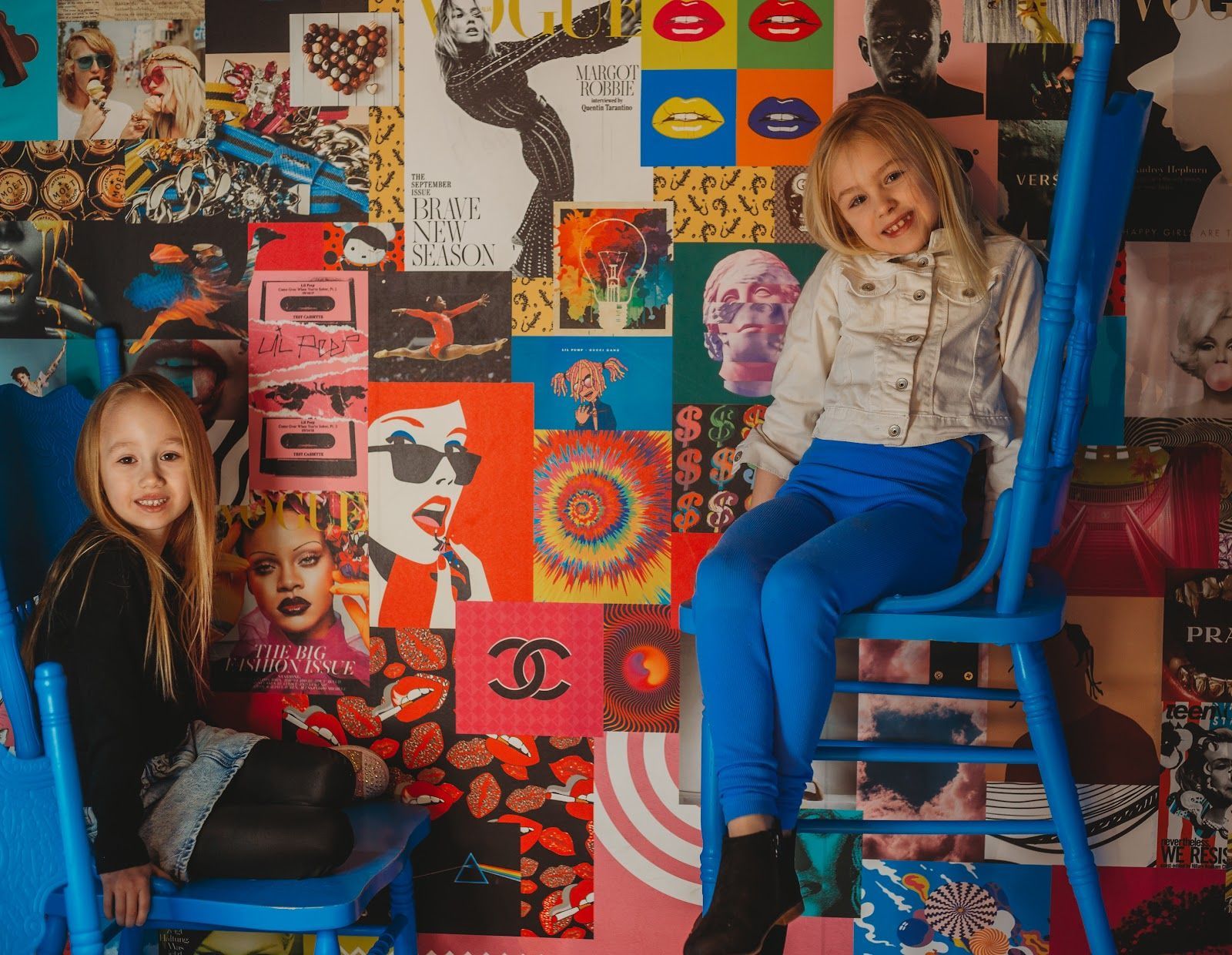 Two little girls are sitting in blue chairs in front of a colorful wall.