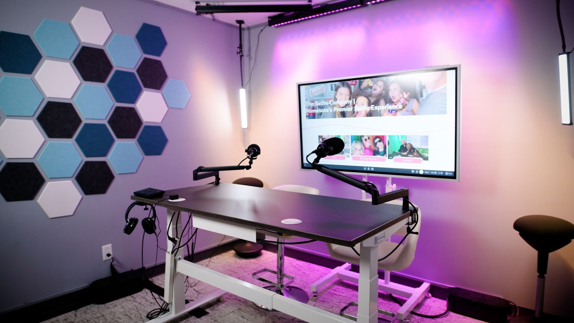 Podcast studio with a desk, mics, and large screen. Hexagon sound panels on the wall.