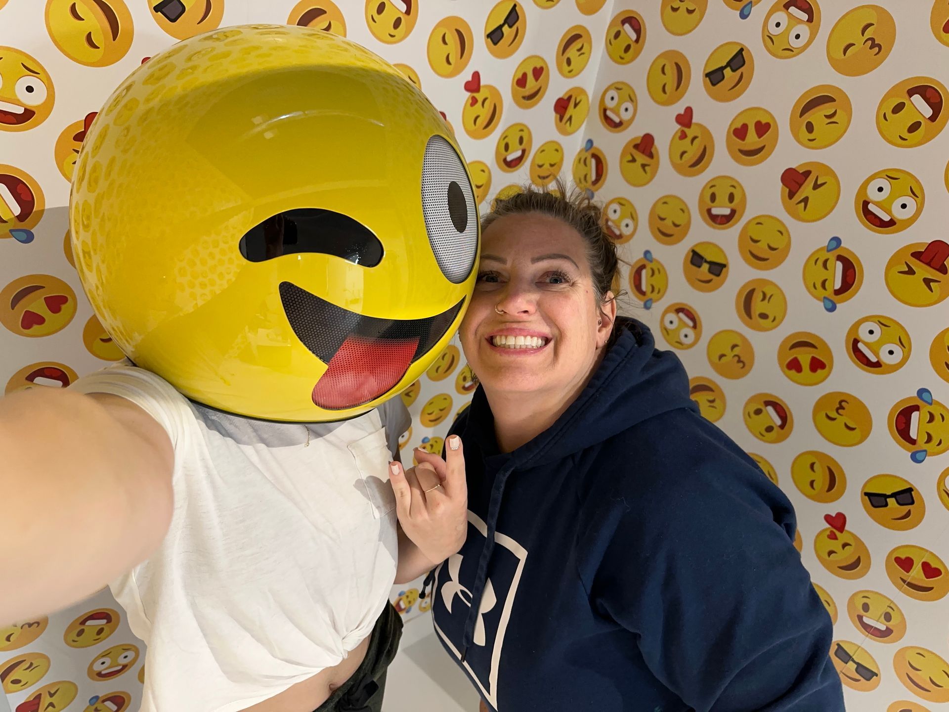 Woman taking selfie with person wearing emoji head, in room with emoji wallpaper.