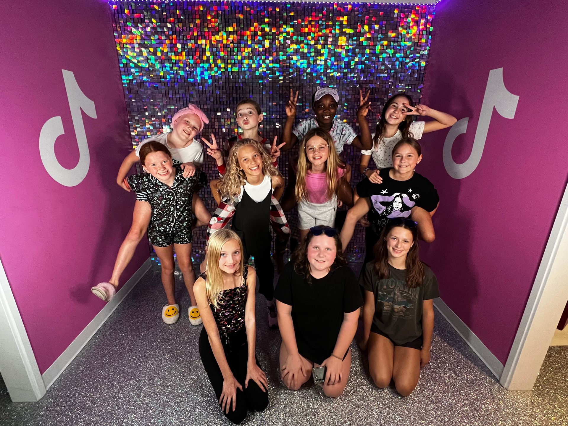 Group of preteen girls posing in a pink room with TikTok logos. Some are making peace signs.