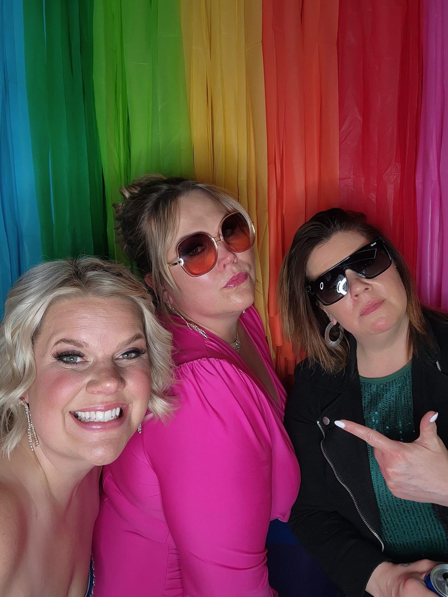 Three women are posing for a picture in front of a rainbow backdrop.