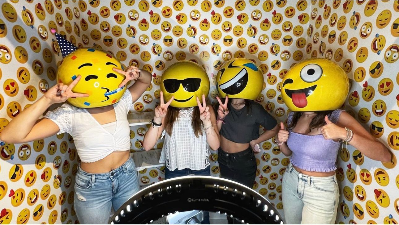 Four young women wearing emoji heads, posing with peace signs and thumbs up, in a room covered with emoji wallpaper.