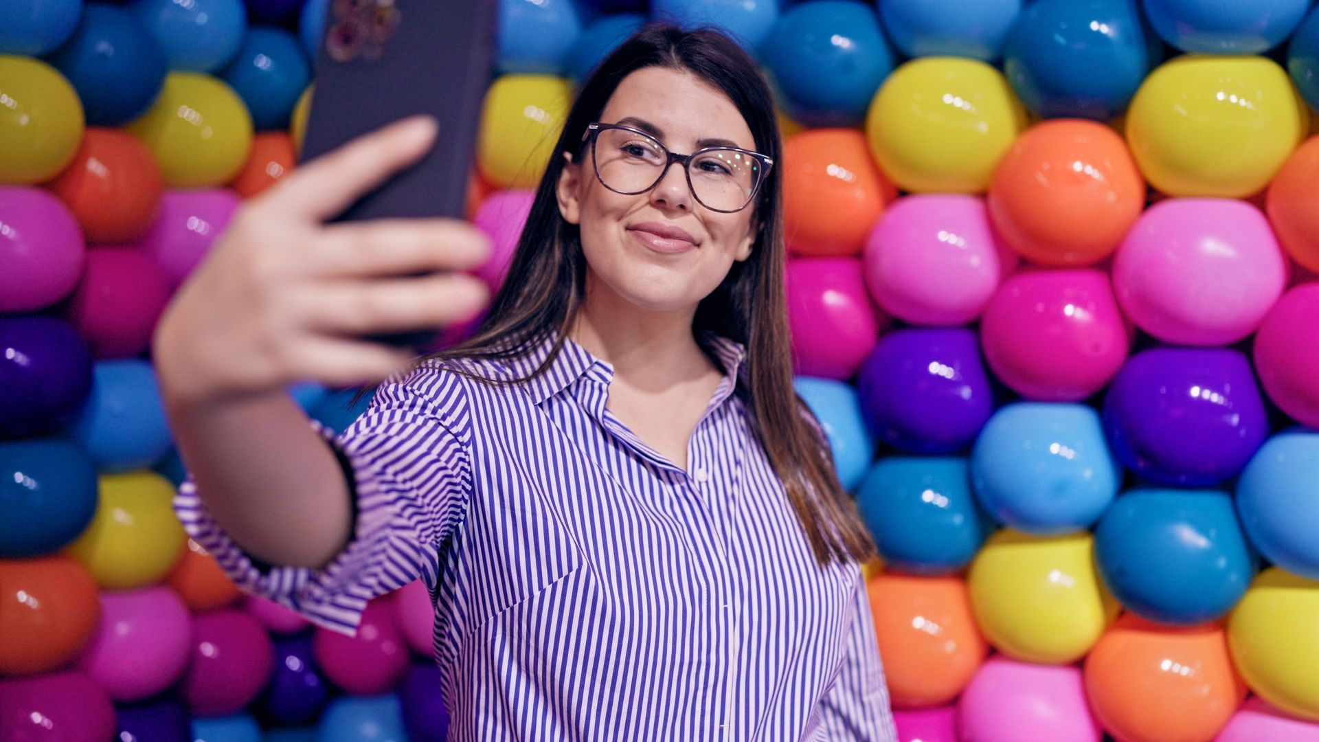 Woman in glasses takes a selfie in front of a wall of colorful balloons.
