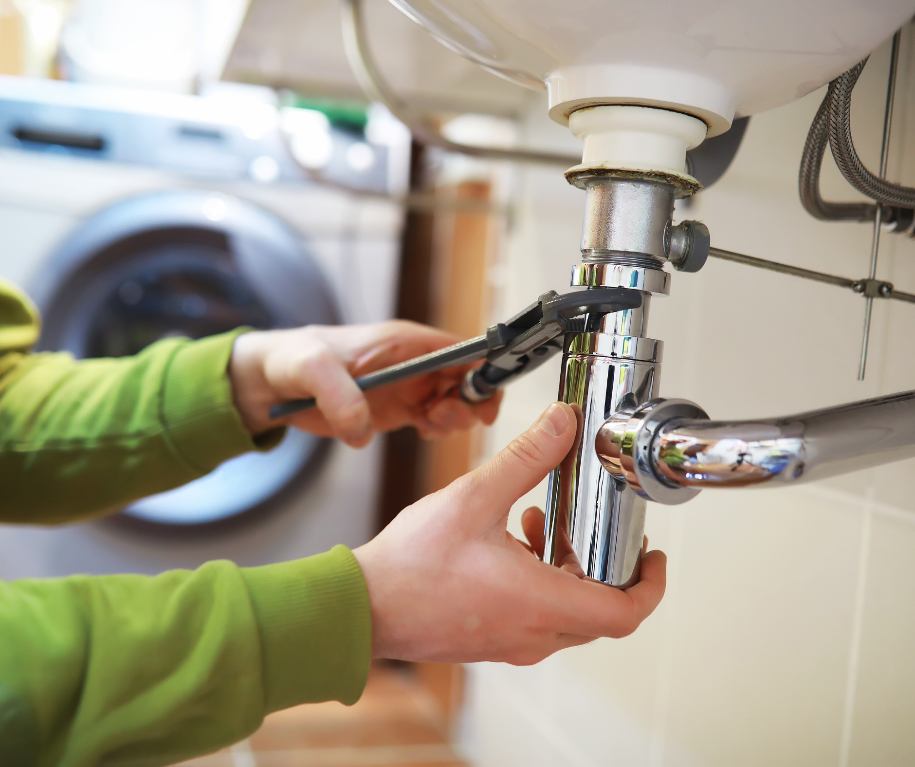 Person using pliers to tighten a chrome pipe under a bathroom sink; washing machine in background.