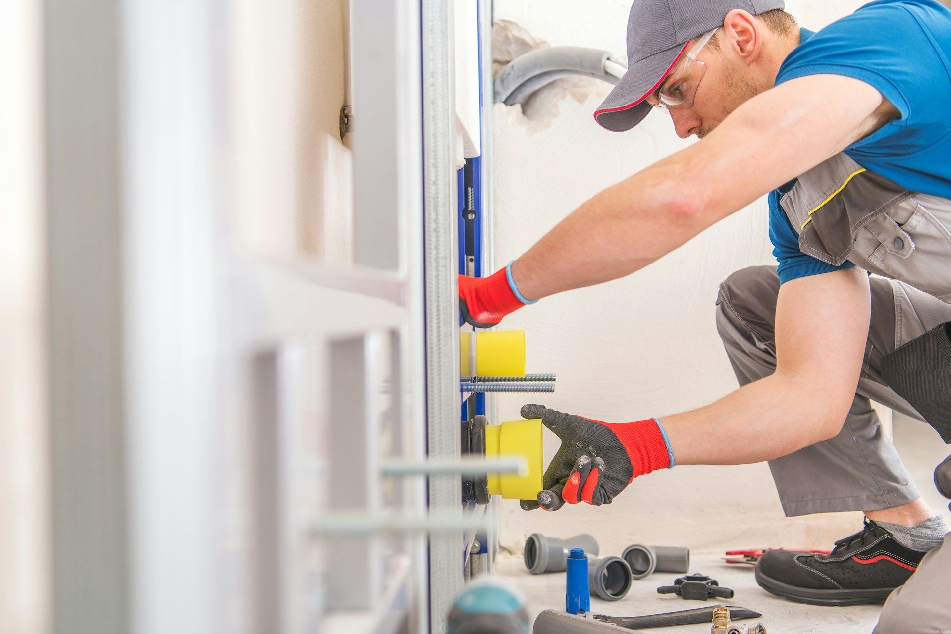 Plumber installing a wall-mounted toilet system; wearing gloves and safety glasses, kneeling, working on the blue frame.