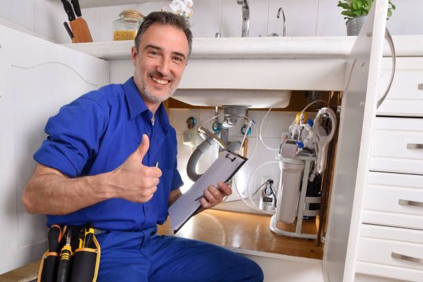 Plumber giving a thumbs up under a kitchen sink. Wearing blue uniform, holding clipboard, and smiling.