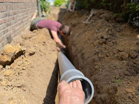 Person installing gray pipe in a trench. Another person works in the background next to a brick wall.