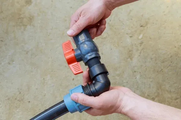 Hands assembling plumbing parts, including a blue connector, black pipe, and red-handled valve.