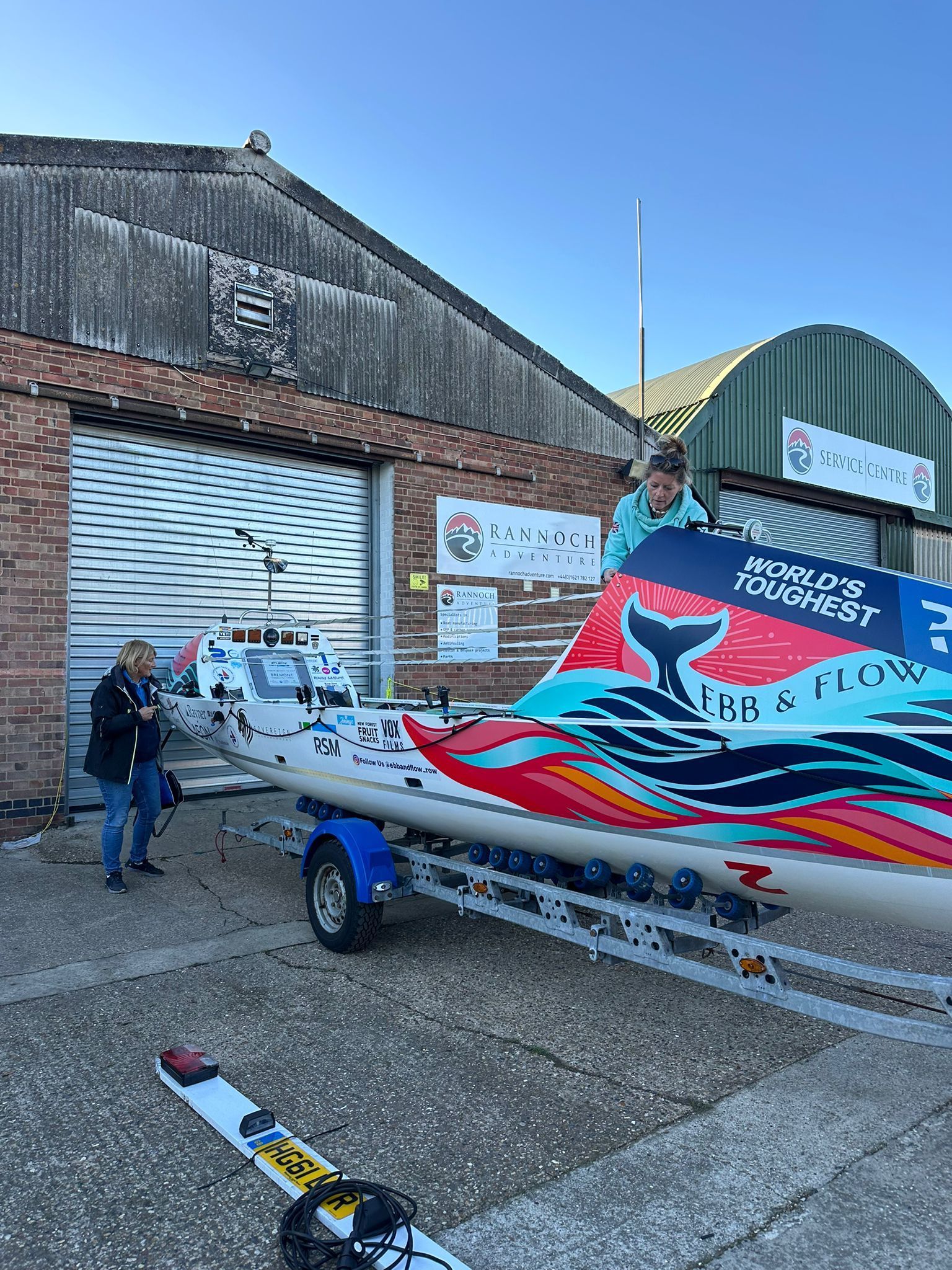 Rowing boat on a trailer, decorated with a whale tail design. Two people stand near the boat outside a building.