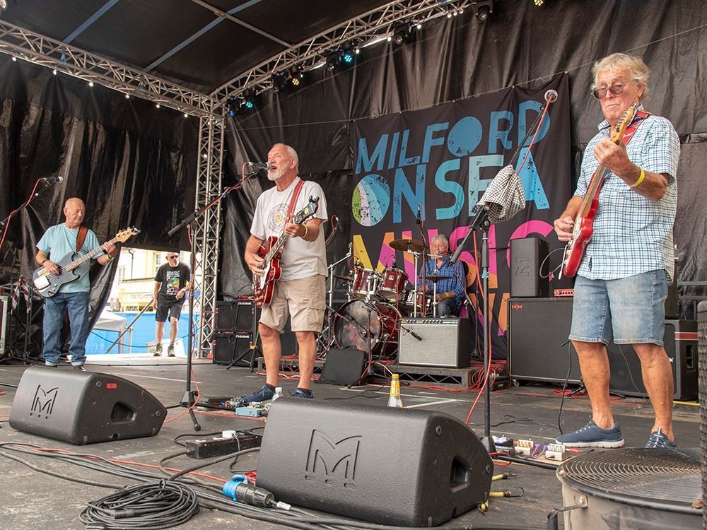 Band performing on outdoor stage at Milford on Sea Music event. Musicians playing guitars, drums. Stage setup with backdrop.