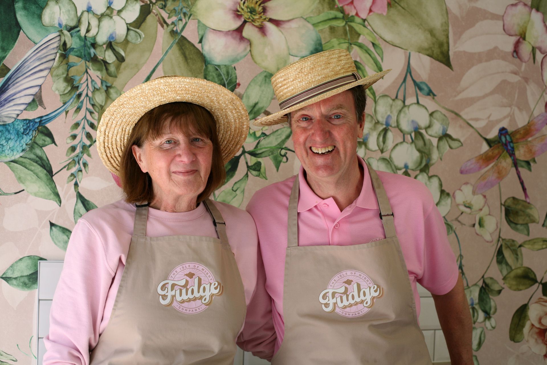 Two people in straw hats and aprons stand in front of floral wallpaper; they are smiling.