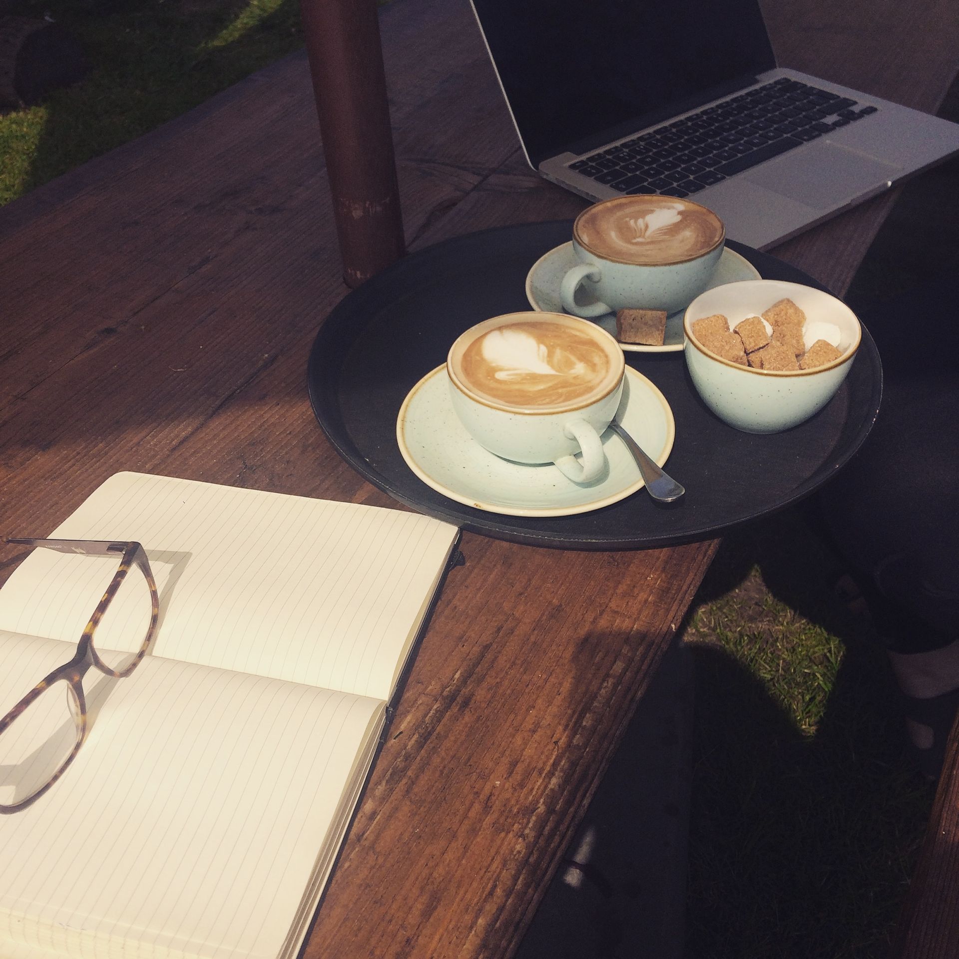Two cappuccinos, cookies, laptop, notebook, and glasses on a wooden table, outside in the sun.