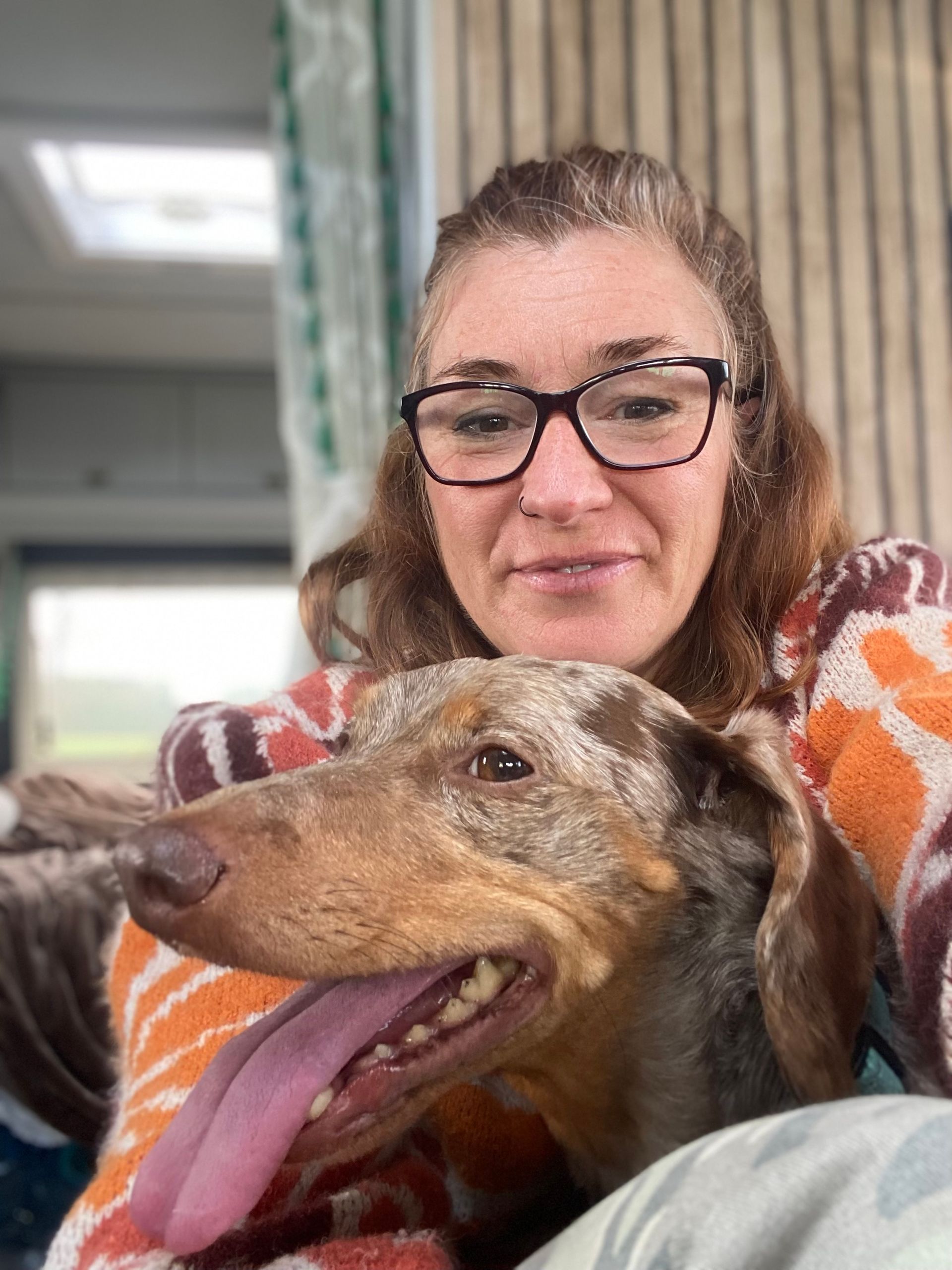 Woman with glasses and dachshund dog, both smiling. The dog's tongue is out. Indoors, natural light.