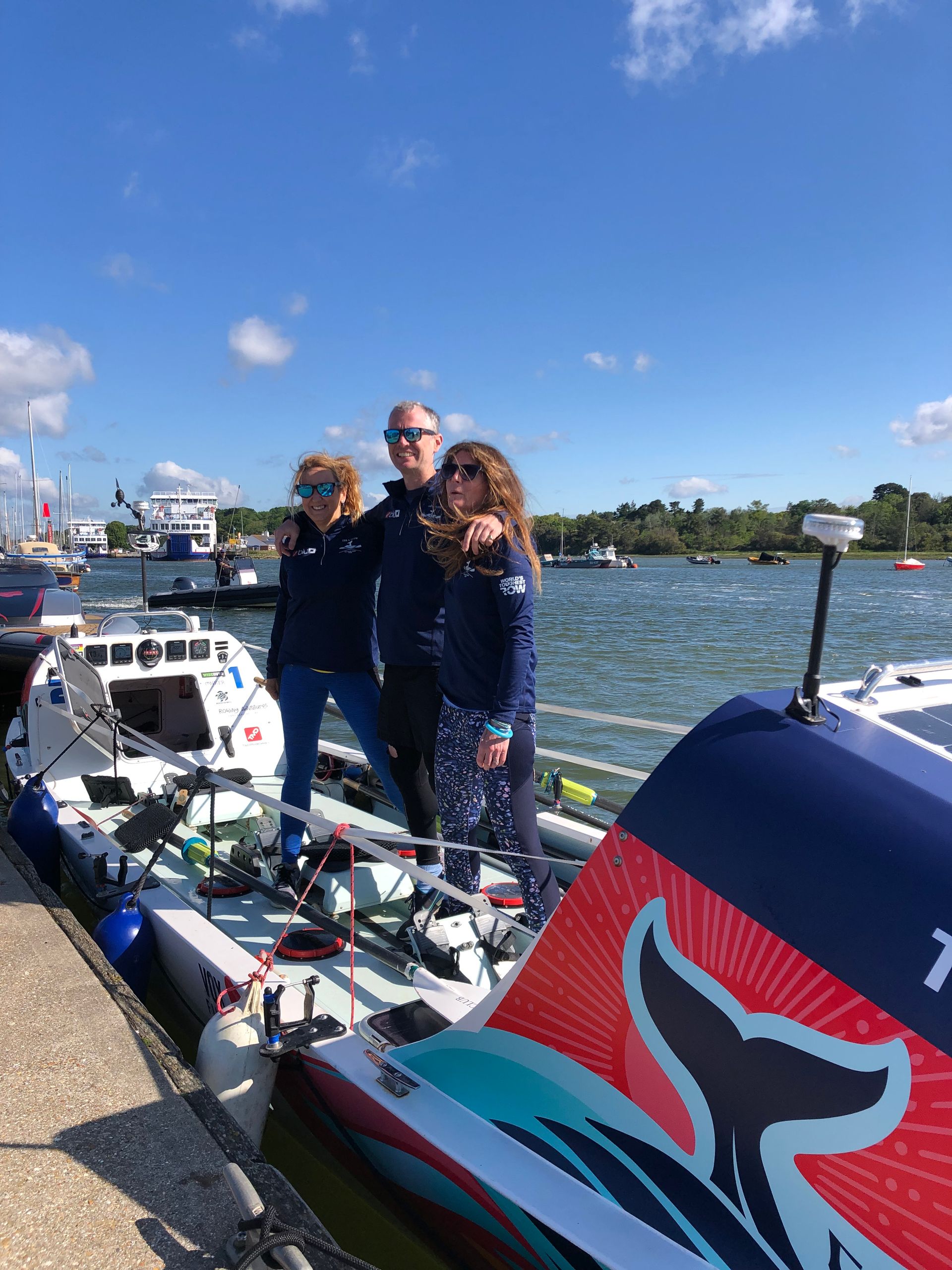 Three people pose by a small boat at a harbor; sunny day, blue tops, boat details, and a whale tail design.