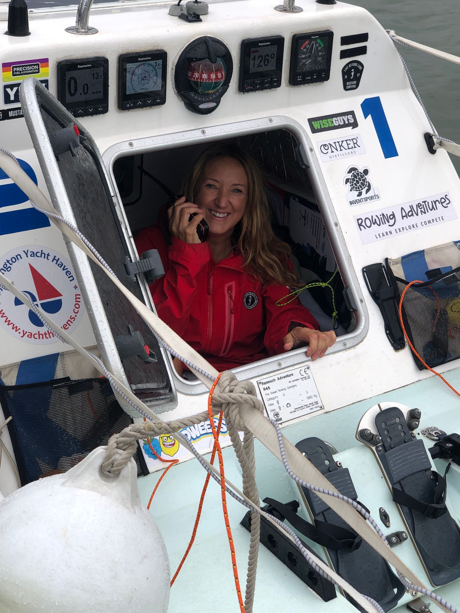 Woman in red jacket smiles while on the phone in a small boat cabin; white and blue setting.