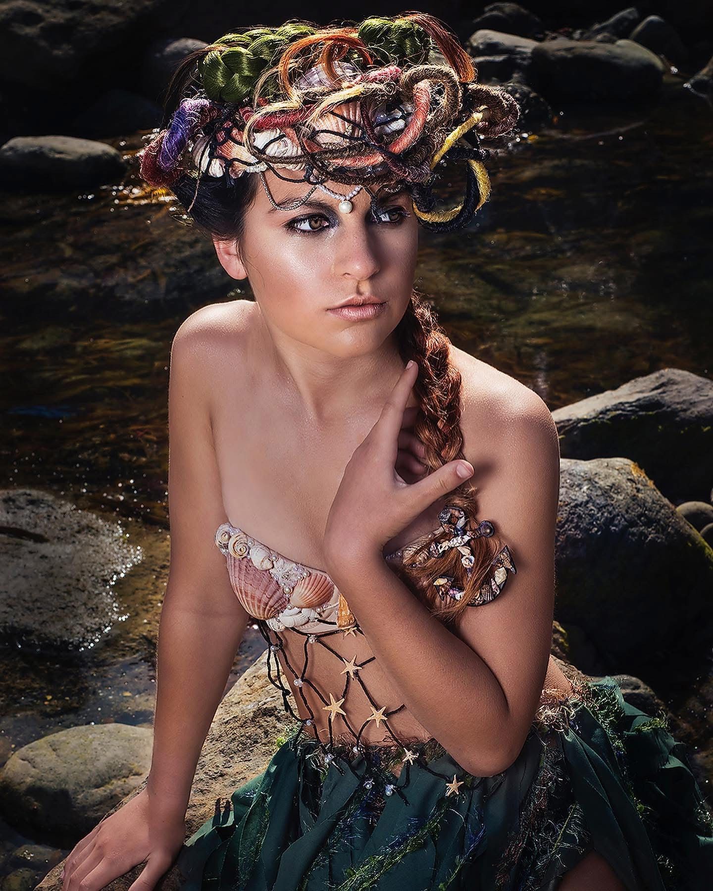 Woman in mermaid-inspired costume, wearing a headpiece, sitting by rocks, looking at the water.