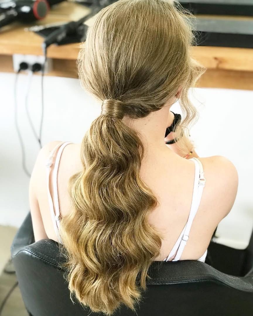 Woman with long, wavy, brown hair in a low ponytail, seated, wearing a white top.