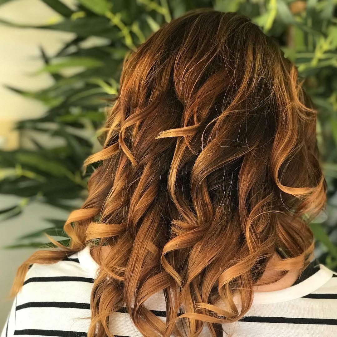 Woman with shoulder-length brown hair styled in loose curls; ombre effect with lighter ends. Green foliage background.