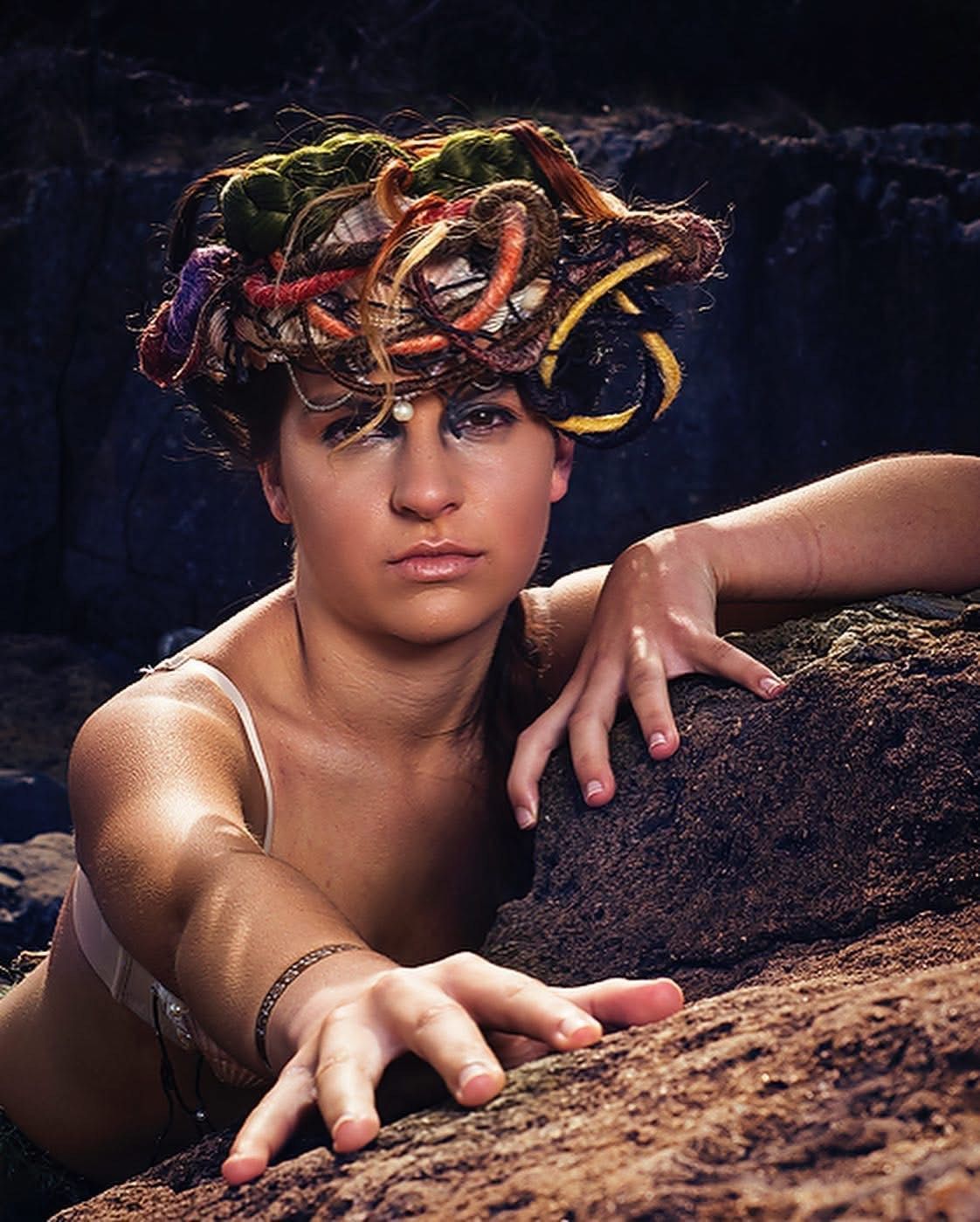 Woman with elaborate hair, reaching out, posing on rocks; ocean background.