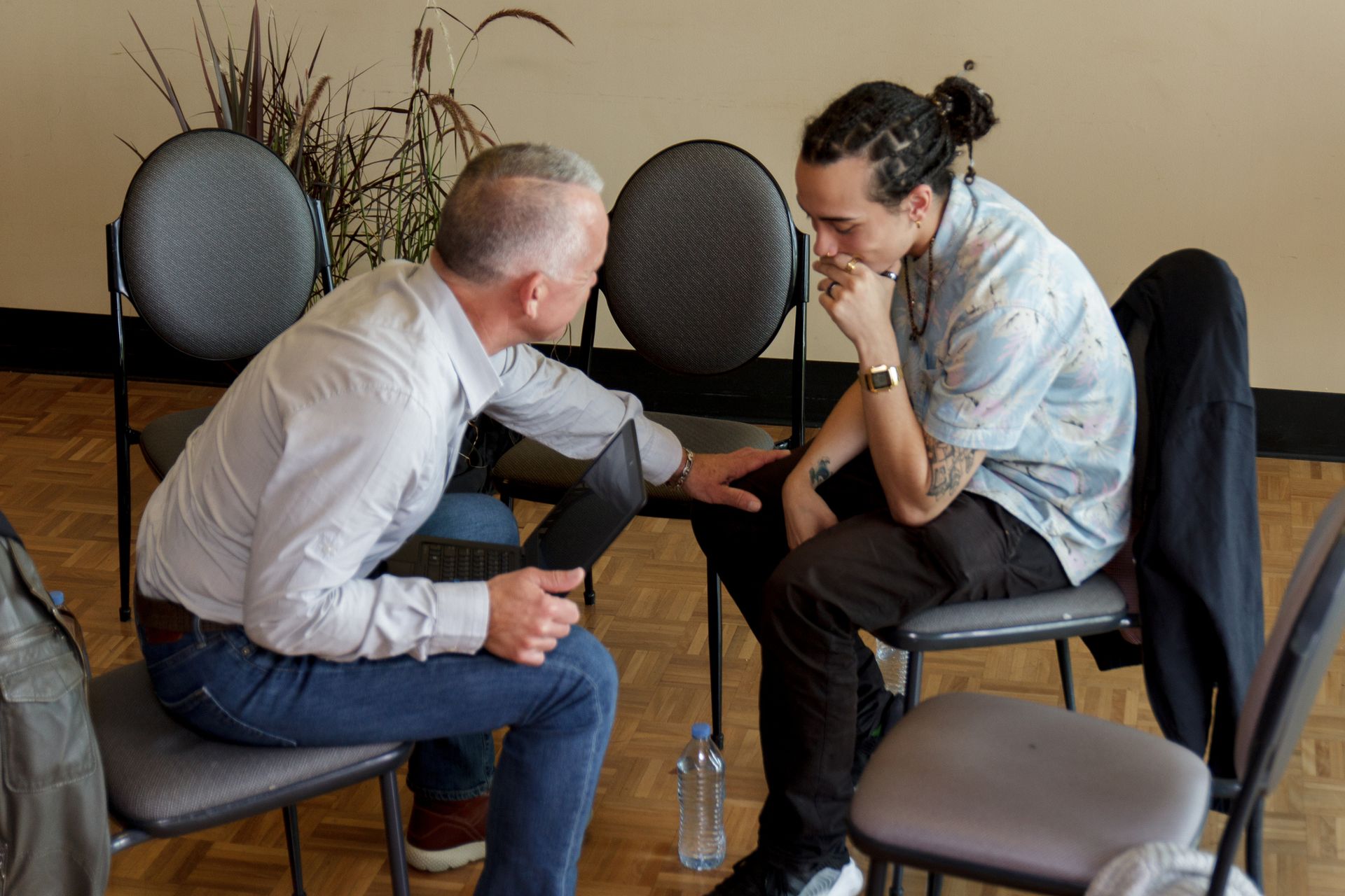 Two men in chairs talking, one pointing at a tablet. Room with chairs, window, and plants.