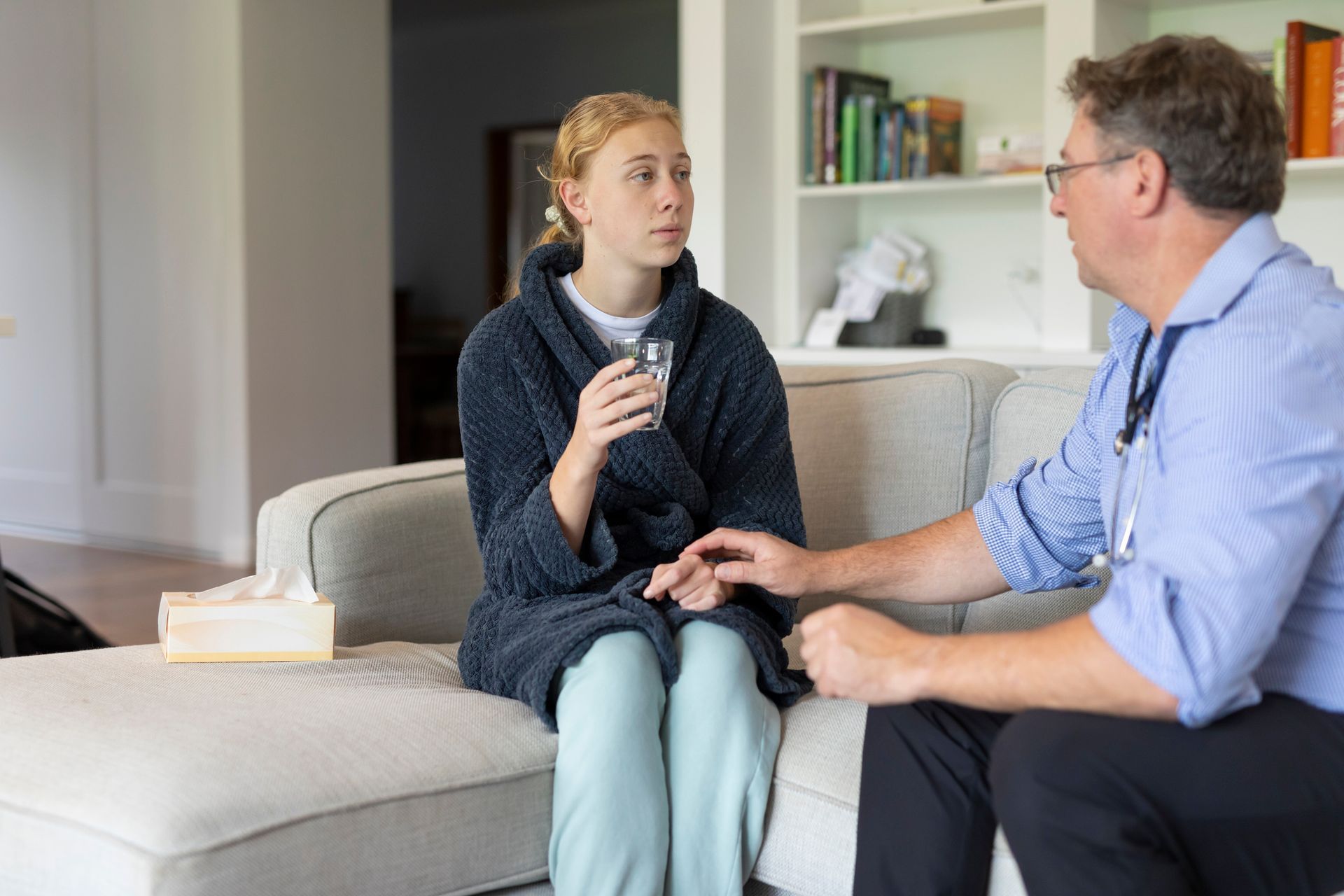 Doctor examining a sick teenage girl on a sofa. Girl in robe holds water glass, doctor has stethoscope.