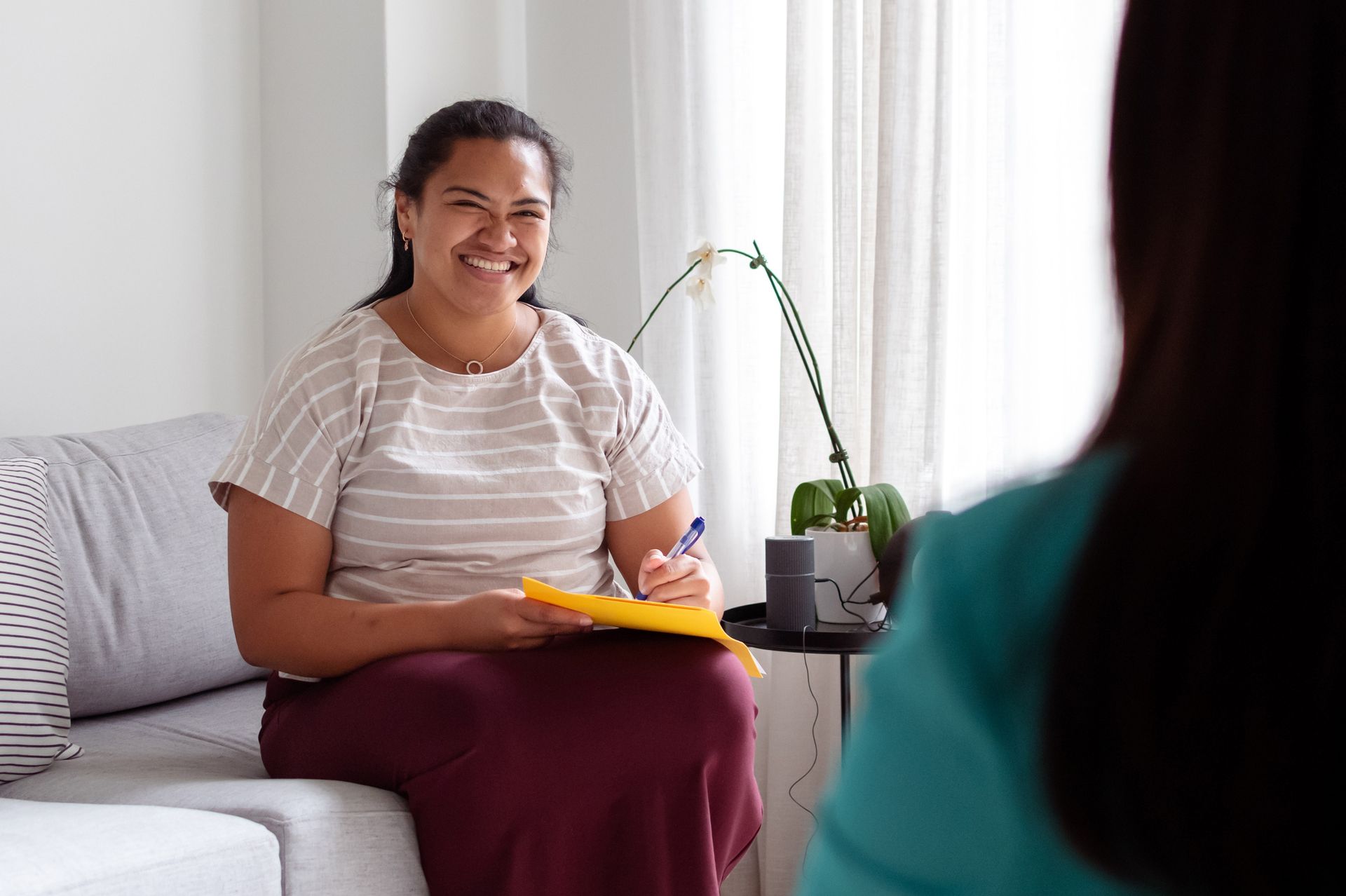 Woman in striped shirt smiles, holding notepad. Sitting on sofa, facing another person in a bright room.