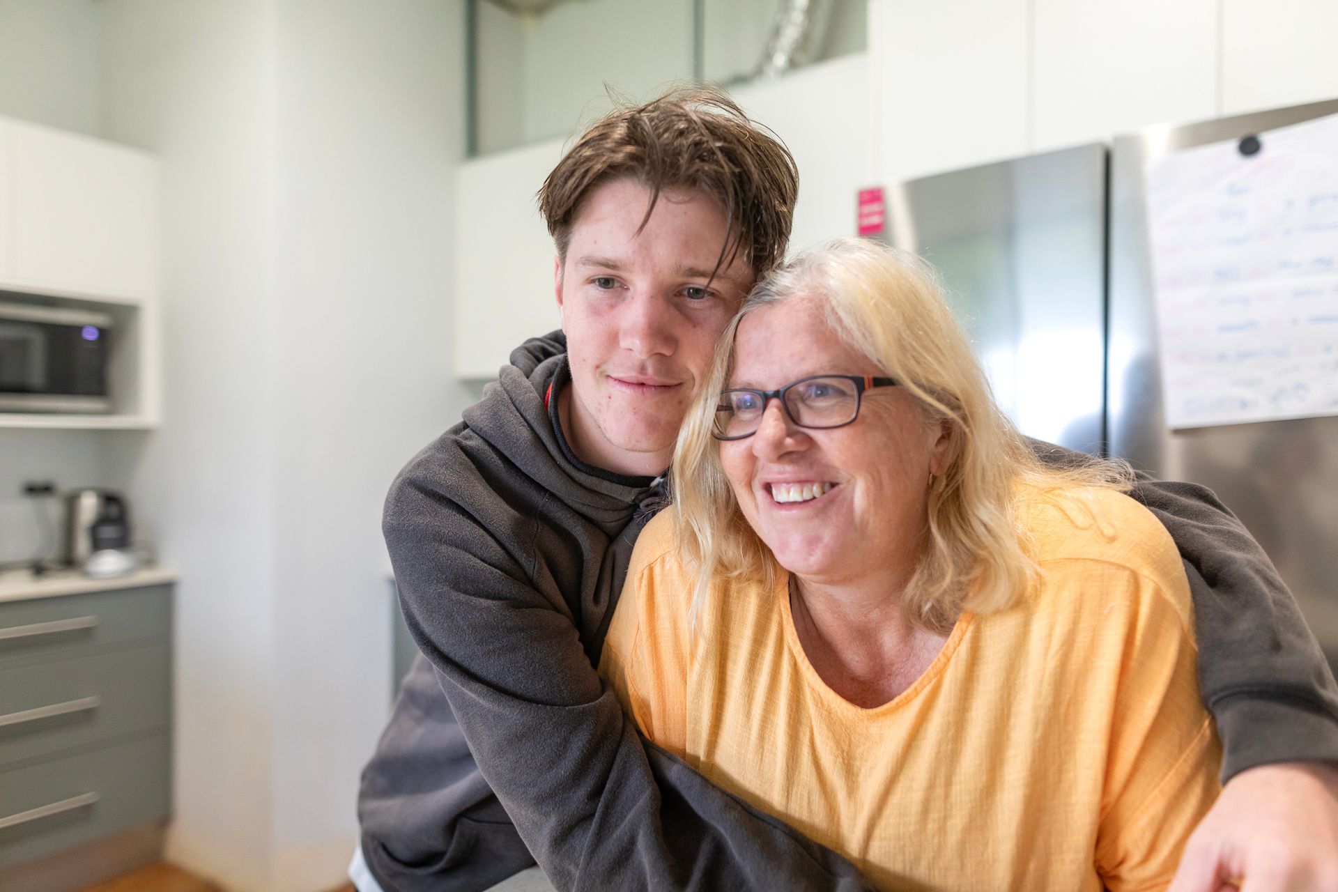 Teen hugs smiling woman in kitchen. She wears glasses, he has brown hair.