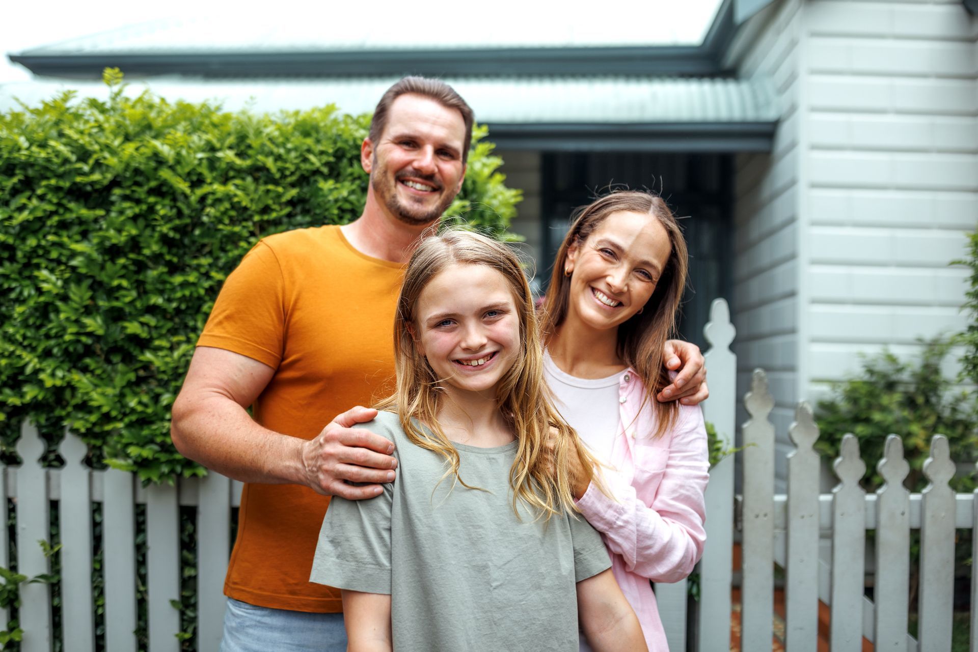 Family of three smiling in front of their house, standing by a white picket fence.