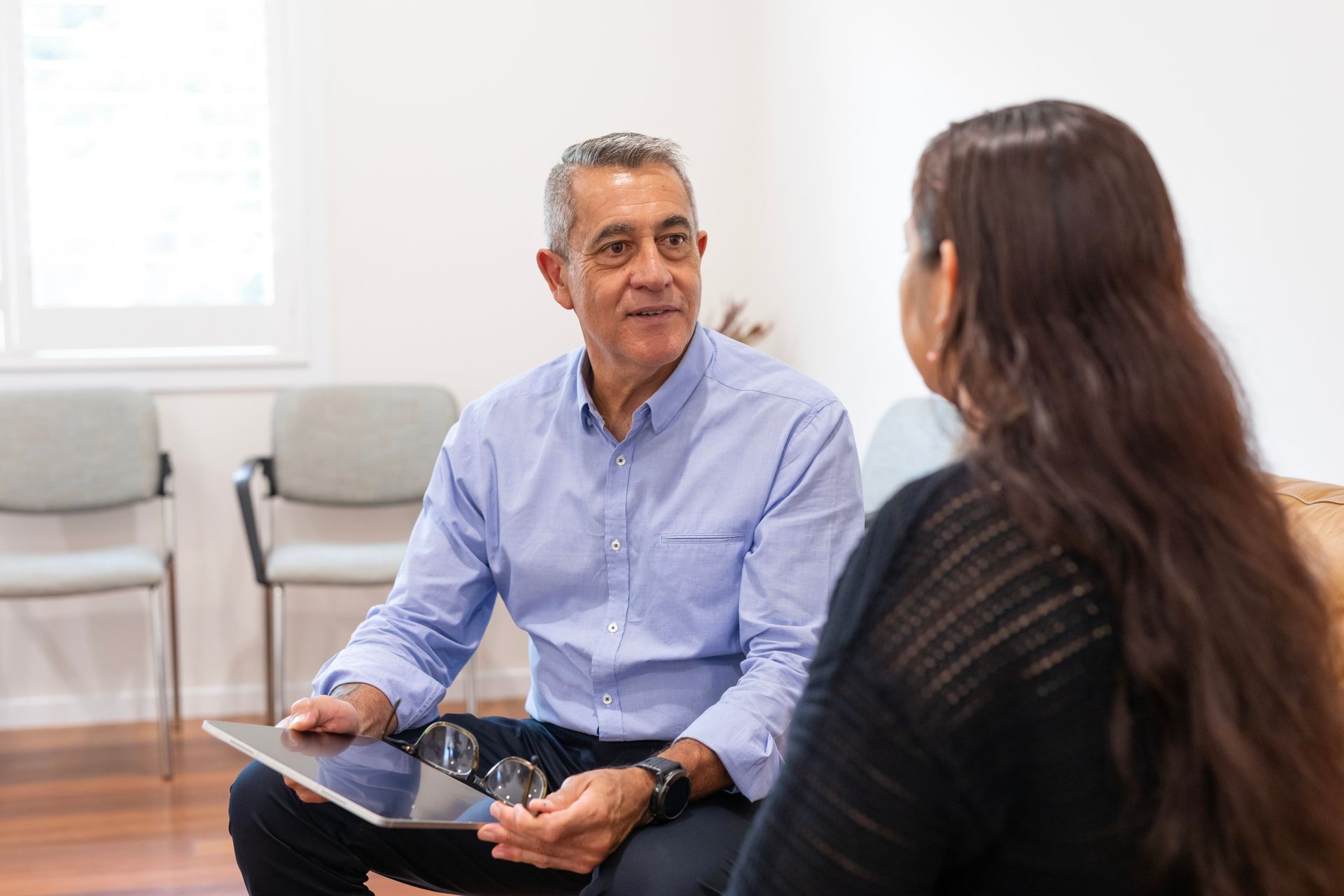 Man in blue shirt and woman conversing, holding tablet and glasses. Sitting indoors.