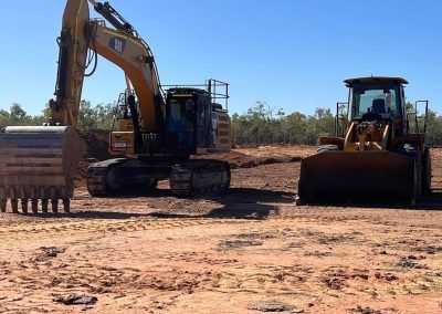 Excavator and front-end loader on dirt ground under blue sky, likely a construction site.