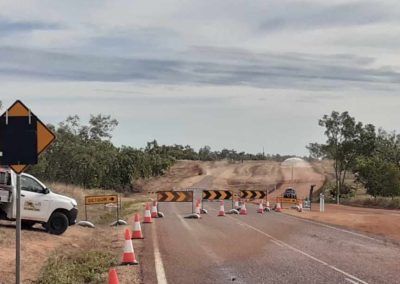 Road closed due to construction. Barricades, cones, and work vehicle block a road. Overcast sky, rural setting.