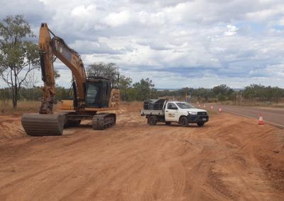 Excavator and white pickup truck on dirt road construction site with cloudy sky.