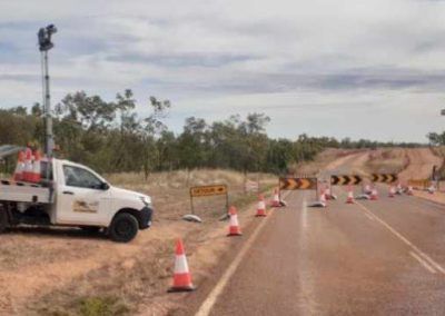 White truck with a tall light tower and orange cones block a road, warning of a curve ahead.