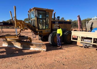 Man standing beside a yellow Caterpillar grader on a dirt road under a clear sky.
